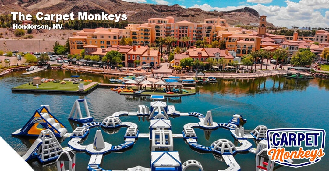 Henderson, NV waterfront view featuring The Carpet Monkeys branding, vibrant buildings, recreational water activities, and inflatable water park structures.